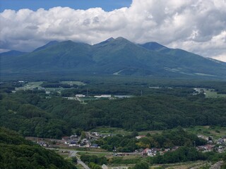 Fototapeta premium Vast mountain range looms above lush green valley. Clouds drift lazily over the peaks. Village sprawls below among trees and fields. Roads wind through quiet countryside