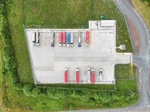 Aerial view of a truck parking lot with several semi-trucks parked in rows on a concrete surface surrounded by green grass and trees in England, United Kingdom.