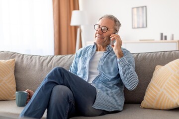 Fototapeta premium Smiling Elderly Caucasian Man Engaged In Phone Conversation While Sitting On Couch At Home, Creating A Comfortable Atmosphere In Modern Living Room, Enjoying Leisure Time And Connection.