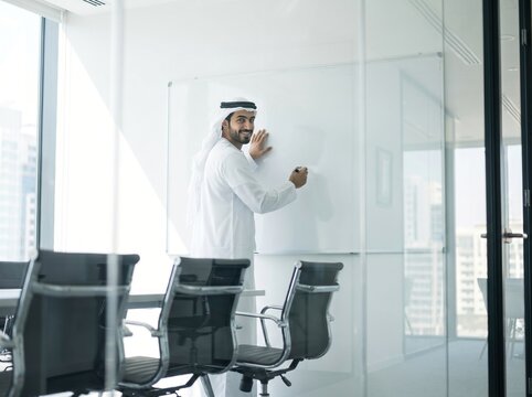 Smiling Arab businessman writing on blank whiteboard in glass meeting room 