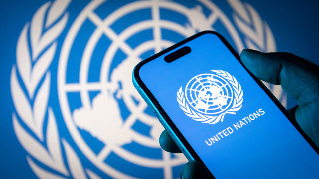 Closeup of a hand holding a smartphone displaying the united nations logo against a blurred un flag background
