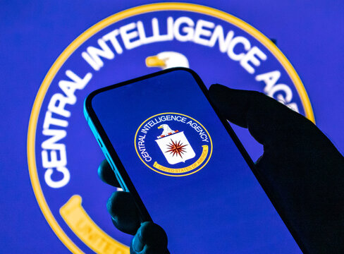 Closeup of a hand holding a smartphone displaying the central intelligence agency logo against a large cia emblem background