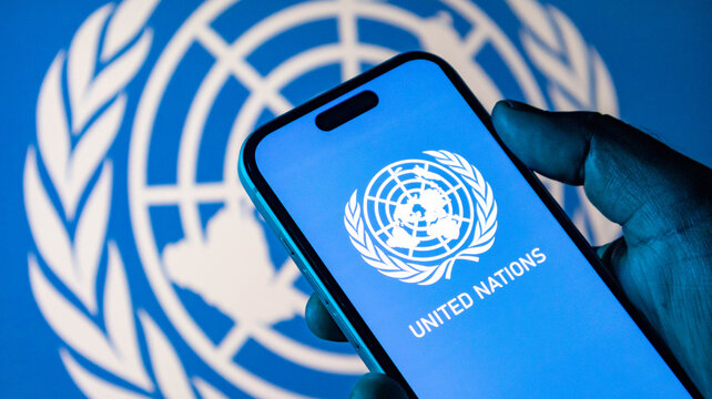 Closeup of a hand holding a smartphone displaying the united nations logo against a blurred un flag background