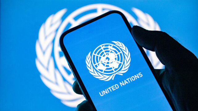 Hand holding a smartphone displaying the united nations logo against a blurred un flag background