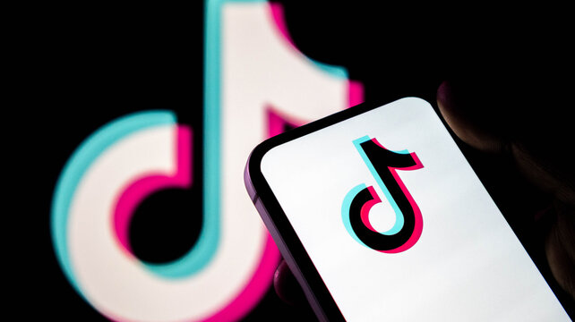 Close up of a hand holding a smartphone displaying the tiktok logo with a large blurred tiktok logo in the background