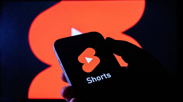 A person holding a smartphone displaying the youtube shorts logo with the same logo in the background