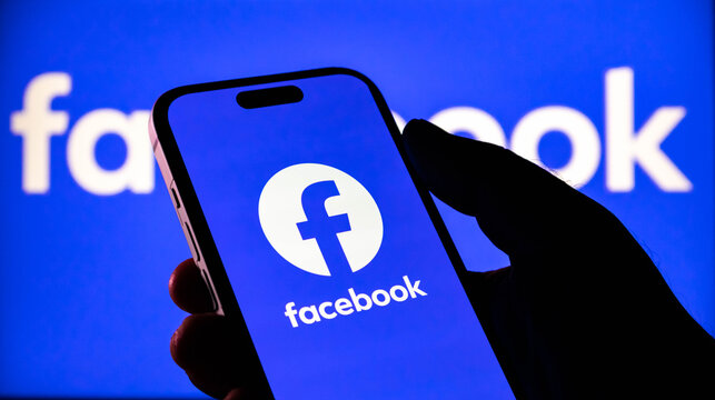 Closeup of a hand holding a smartphone displaying the facebook logo with the facebook logo in the background