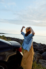 Fototapeta premium A woman stands beside a car on a rocky coast, arms raised in celebration, enjoying fresh air and sunlight. A candid lifestyle moment outdoors.