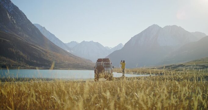 Traveler in a yellow jacket taking a smartphone photo near an off road SUV parked by a mountain lake in a remote alpine valley. Golden grass, snowy peaks, and bright sunlight create a scenic outdoor