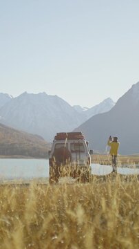 Traveler in a yellow jacket taking a smartphone photo near an off road SUV parked by a mountain lake in a remote alpine valley. Golden grass, snowy peaks, and bright sunlight create a scenic outdoor