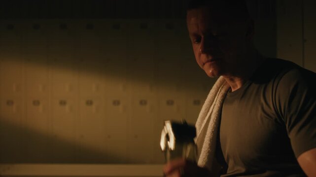 Cinematic Close Up Man Heavy Breathing and Determination in Warm, Atmospheric Light of Locker Room. Young Man With Short Hair, Covered in Sweat, Wipes Face With Towel, Drinks Water from Sports Bottle.