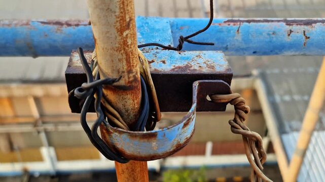 Rusty metal clamp with bolt holding weathered industrial wires on steel pipe scaffolding.
