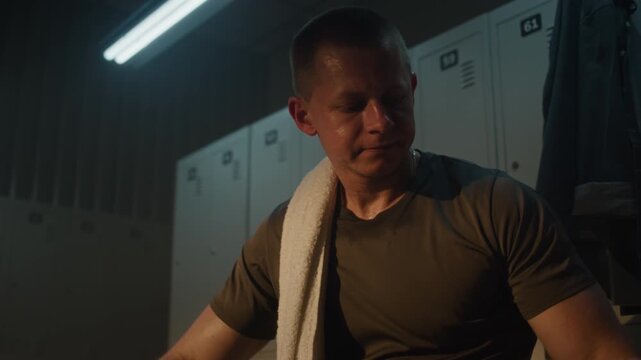 Athlete in Warm Lit Locker Room, Catching Breath After Intense Workout. Sweat Glistens on Face as Man Wipes With Towel, Takes Deep Drink from Water Bottle. Raw Effort and Determination in Recovery.
