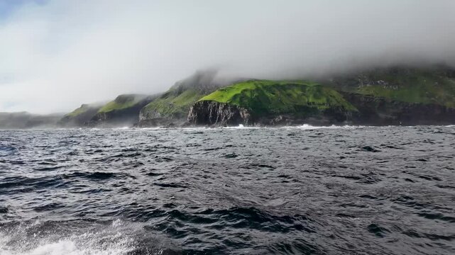 Atmospheric water view of lush green coastal cliffs of a neighboring island partially covered in mist and low clouds near Mykines, Faroe Islands - 안개와 낮은 구름에 부분적으로 뒤덮인 푸른 해안 절벽과 페로 제도 미키네스 주변 섬의 선상 바다