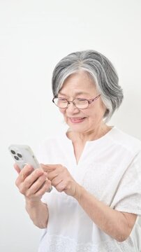 白背景でスマートフォンを見る操作、タップする笑顔のグレイヘアのシニア女性 登録や買い物などの縦動画