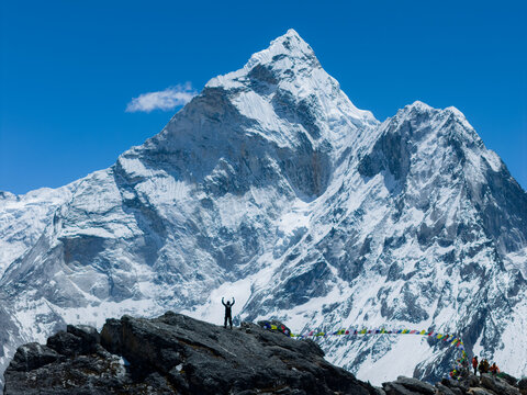 Aerial view of Ama Dablam peak with snow-covered slopes and rocky terrain featuring a climber standing with arms raised and colorful prayer flags in Nepal.