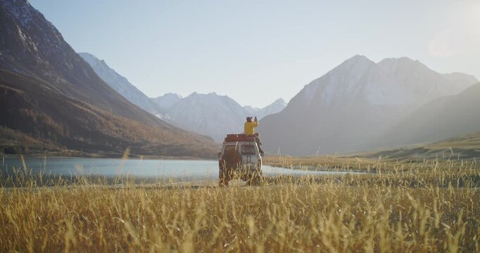 Man in a yellow jacket sitting on the roof of an off road vehicle and taking a phone picture near a mountain lake with snowy peaks in the background. Warm sunlight, golden grass, and open wilderness