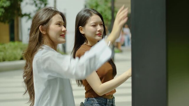 Two beautiful young Asian women cheerfully using an outdoor interactive digital touch screen kiosk in the city. They are smiling, talking, and pointing at the directory to find helpful info.