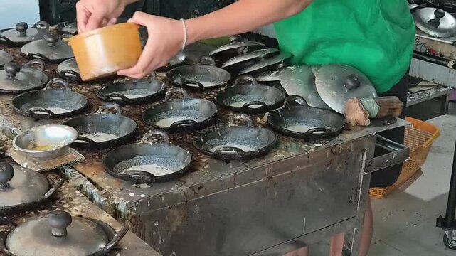 Close-up of a vendor pouring rice flour and coconut milk batter into multiple small hot woks with a ladle while preparing traditional Indonesian Surabi Solo cakes.