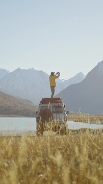 Man in a yellow jacket standing on the roof of an off road vehicle and shooting a phone picture by a mountain lake with snowy peaks in the background. Wide natural landscape with dry grass and bright