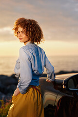 Fototapeta premium A woman stands beside a car at sunset, embracing a casual outdoor lifestyle with a confident pose, highlighting fashion and a striking portrait, in warm light.