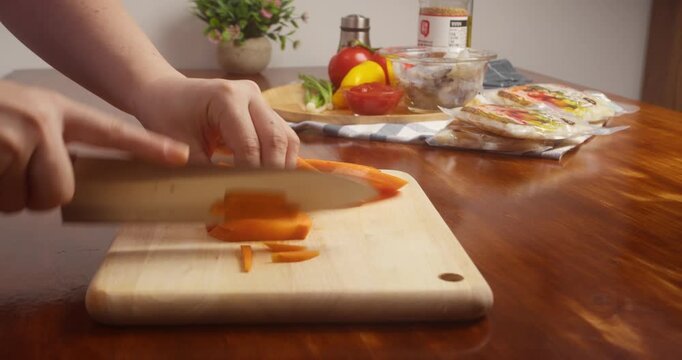 Close up of hands slicing fresh carrot into thin strips with a kitchen knife on a wooden cutting board during vegetable preparation for home cooking.