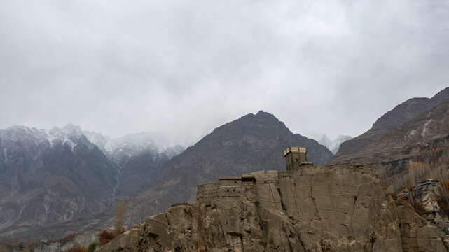 Aerial view of Altit Fort perched on a rocky cliff with snow-capped mountains in the background under a cloudy sky in Altit, Gilgit, Pakistan.