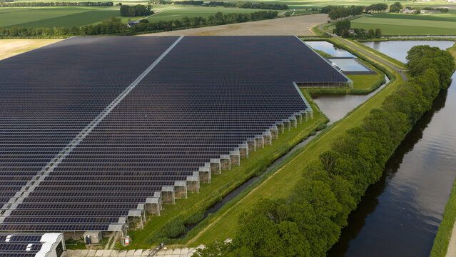 Aerial view of a large solar panel farm integrated with greenhouse structures, situated next to a canal and surrounded by agricultural fields in Middenmeer, North Holland, Netherlands.