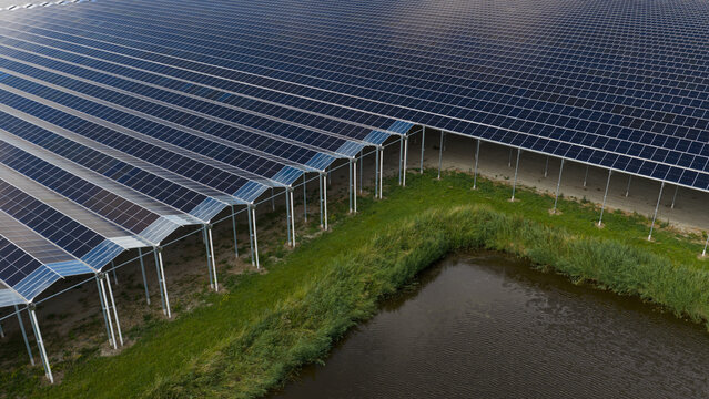 Aerial view of a large solar panel array installed on an elevated structure above a green field and a small pond in Middenmeer, North Holland, Netherlands.