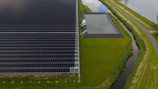 Aerial view of a large solar panel array installed on a greenhouse roof next to a water reservoir and a canal in the rural landscape of Middenmeer, North Holland, Netherlands.