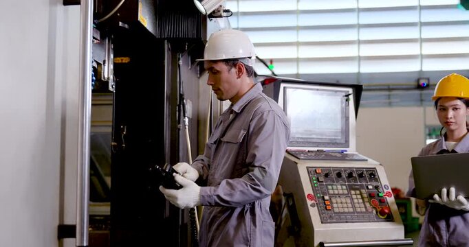 Asian man engineer examines CNC machine area while Asian woman technician reads laptop data, coordinating process verification and connected production monitoring in manufacturing plant, slow motion