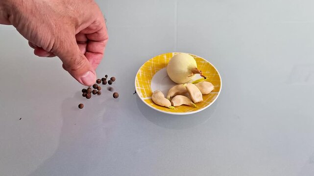 Whole onion and garlic cloves arranged on a plate with scattered peppercorns, presenting fresh cooking ingredients in a simple kitchen food preparation and seasoning scene