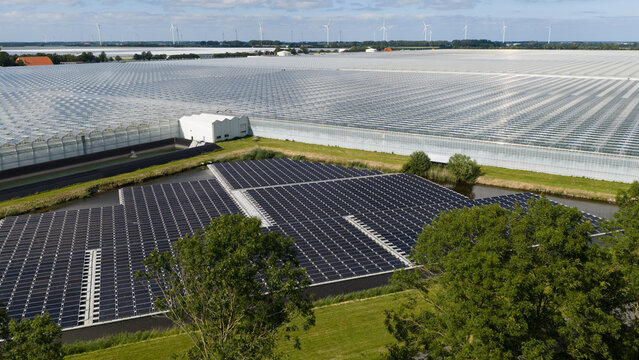 Aerial view of a large greenhouse complex and a floating solar panel array installed on a canal in Middenmeer, North Holland, Netherlands.