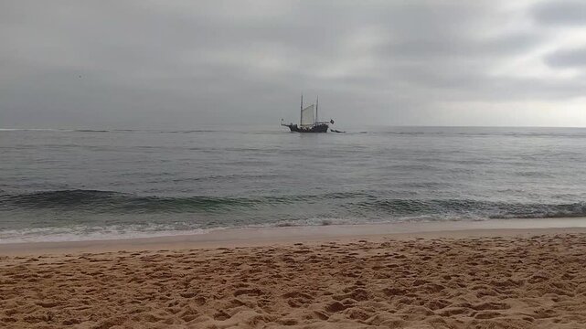 Sailing boat moves slowly on the water near the beach during cloudy weather in a coastal area