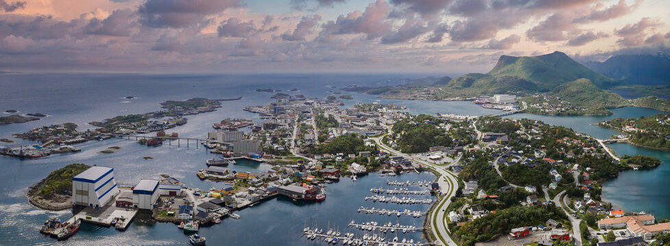 Aerial view of Svolvaer, Lofoten, Norway, with marinas, clustered fishing harbors, blue roof shipyards, curved roads, and jagged mountains under soft evening light.
