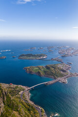 Obraz premium Aerial view of Lofoten, Norway, shows a curving bridge over turquoise water toward rocky islets and fishing settlements near Henningsvaer in clear daylight.