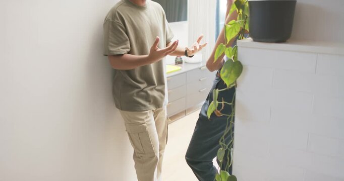 Couple catching up in kitchen after man shifting weight on wall, woman listening near pothos