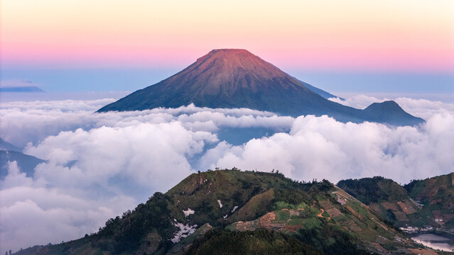 Aerial view of Mount Sindoro rising above a thick sea of clouds with terraced hills and a colorful sunset sky in Indonesia.