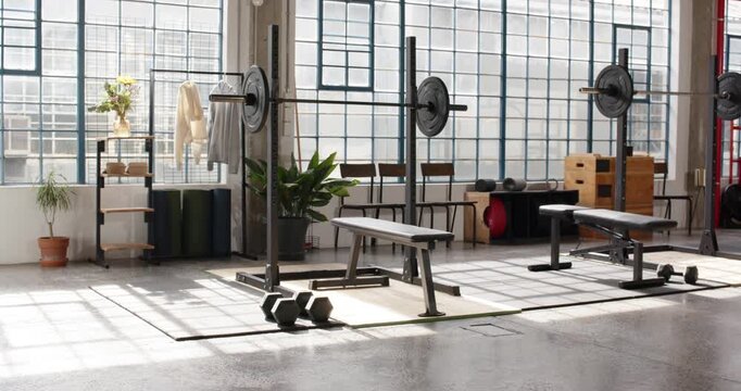 Sunlight is moving through windows casting shadows across bench, barbell and dumbbells in loft gym