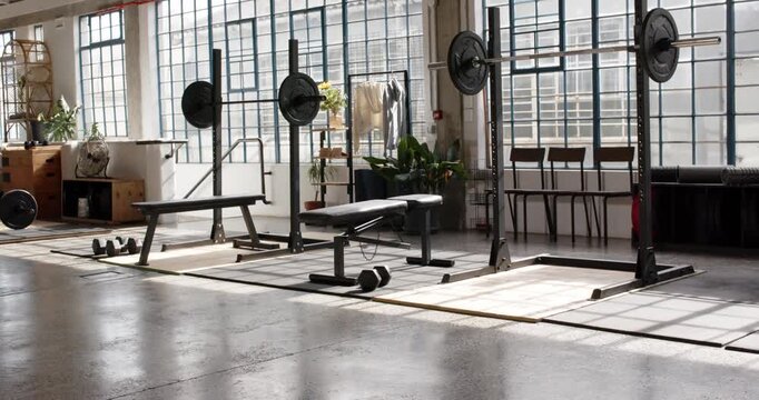 Camera is panning right, framing bench and squat racks while sunlight is shifting, showing loft gym