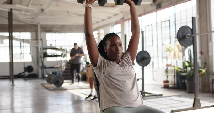 African woman pressing hex weights overhead on incline bench from bent elbows for shoulder strength