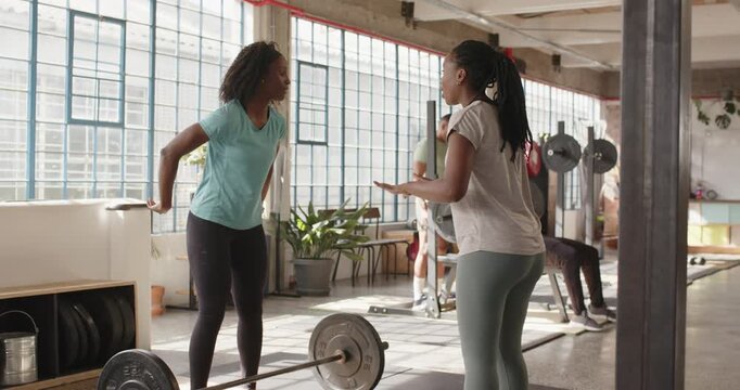 African American women trainer cueing trainee, trainee gripping barbell and lifting for form in gym