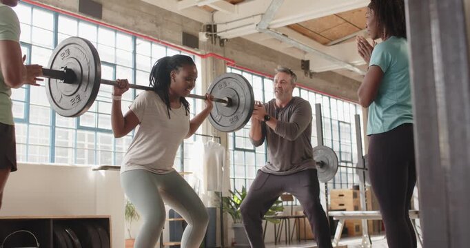 Diverse group trainer urging African American woman squatting with loaded bar for rep at loft gym
