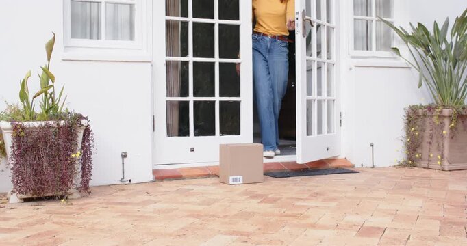 Adult woman in mustard top noticing package, opening white doors, stepping onto patio, lifting box