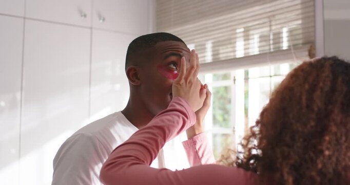 Couple by window blinds, woman reaching, applying pink undereye gel patches for care, man laughing