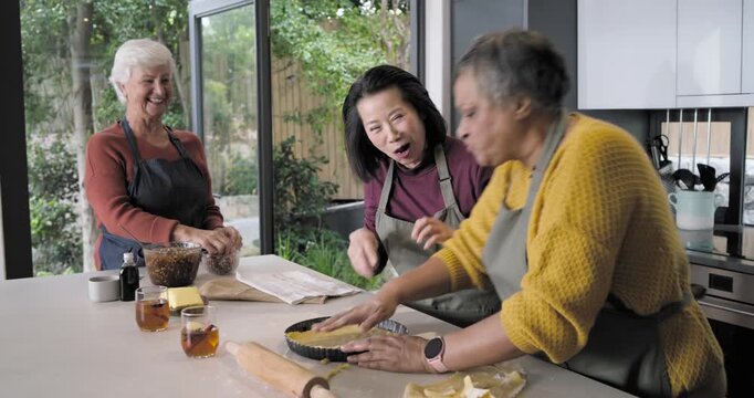 Diverse women in aprons using recipe, rolling dough, pressing crust into tart pan at kitchen island
