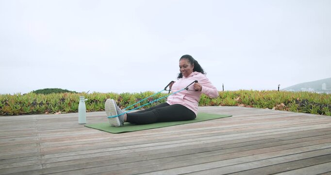 Senior African-American woman pulling band anchored around feet on mat on deck for fitness bottle