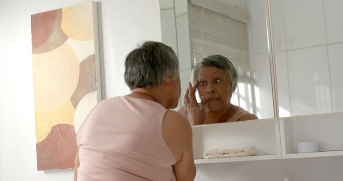 Noticing under-eye Senior African American woman dabbing and checking face at vanity near mirror