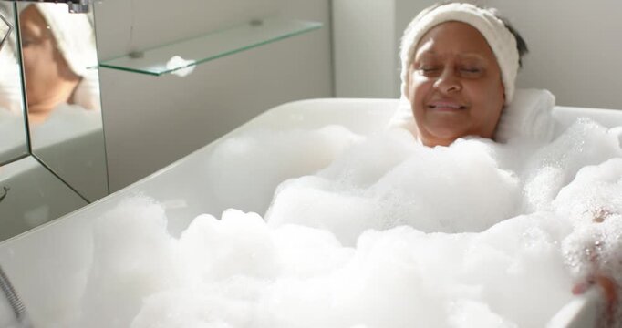 African American woman resting in bathroom tub amid foam rolled towel headband camera pulling back