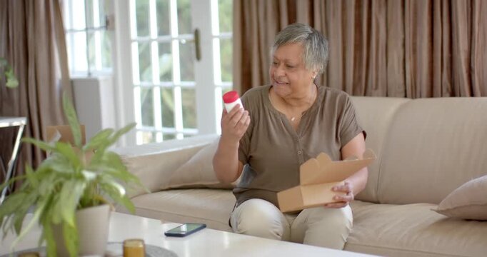 African American woman opening parcel on couch, examining white bottle red cap, confirming contents